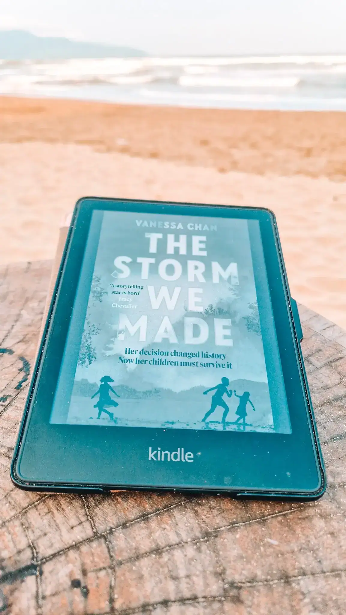 A Kindle with the cover for 'The Storm We Made' sits on a wooden table with sand visible on a beach.