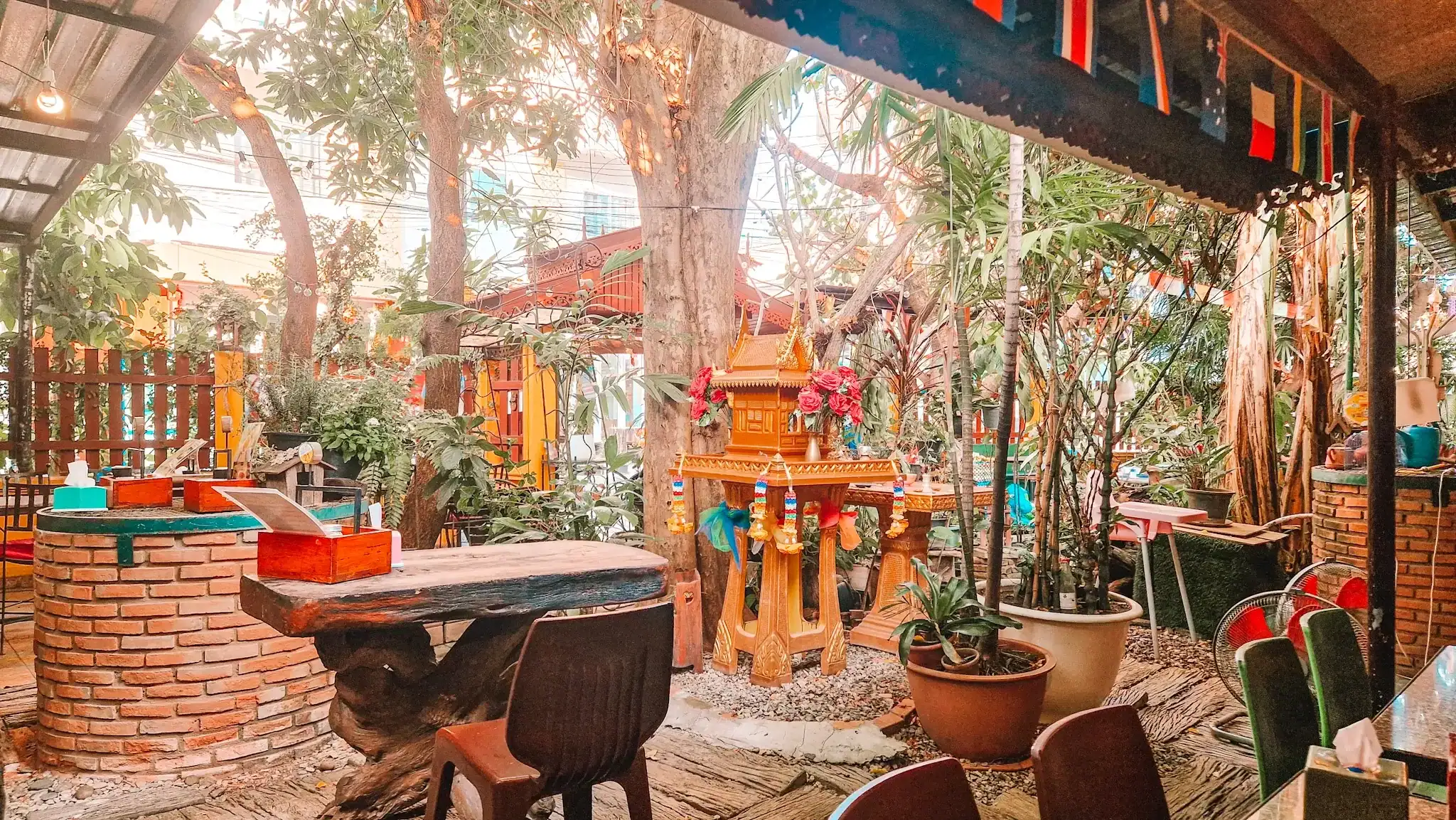 A vibrant garden dining area with a mix of wooden and brick furniture, centered around a golden Thai spirit house adorned with garlands and surrounded by greenery and colourful flags, at Blue Diamond Cafe, one of the best coffee shops in Chiang Mai