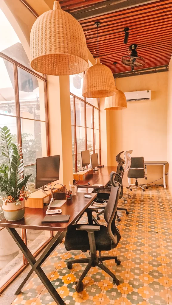 A light and airy office space, with big windows broken up into smaller panes. Desks line the windows wtih monitors on riders and ergonomic chairs in front of each desk. There is a green plant in the foreground and a tablet and stand on the nearest desk.
