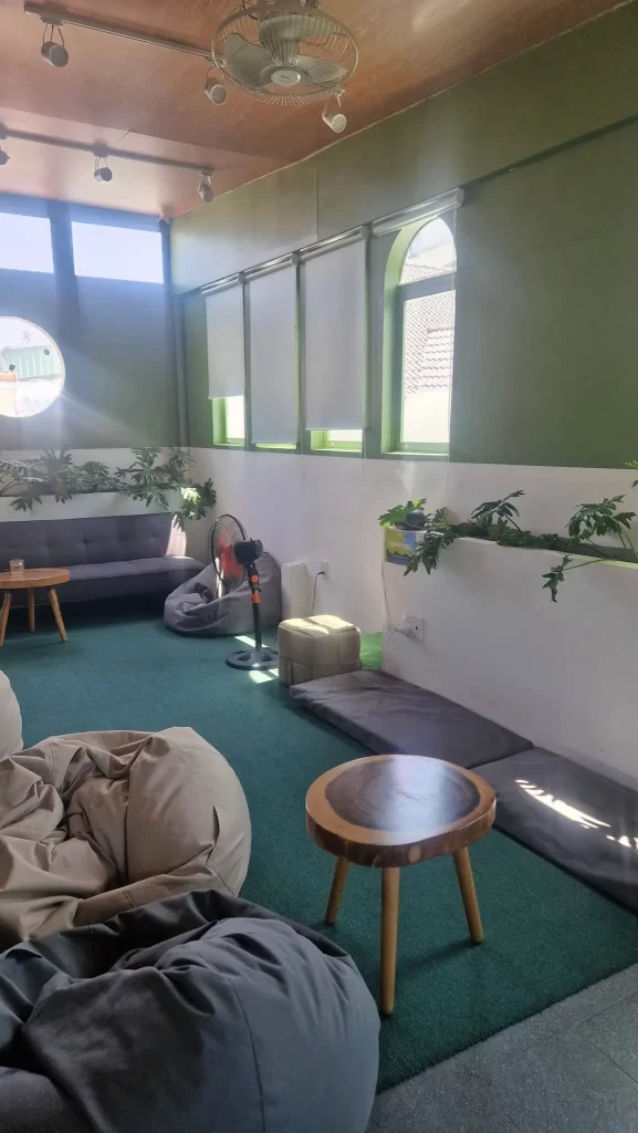A relaxing area with bean bags, floor cushions, a sofa and side tables, all in shades of grey and green. There are plants dotted around and sunlight coming through the windows. There are fans on the ceiling and a floor fan pointing at the sofa