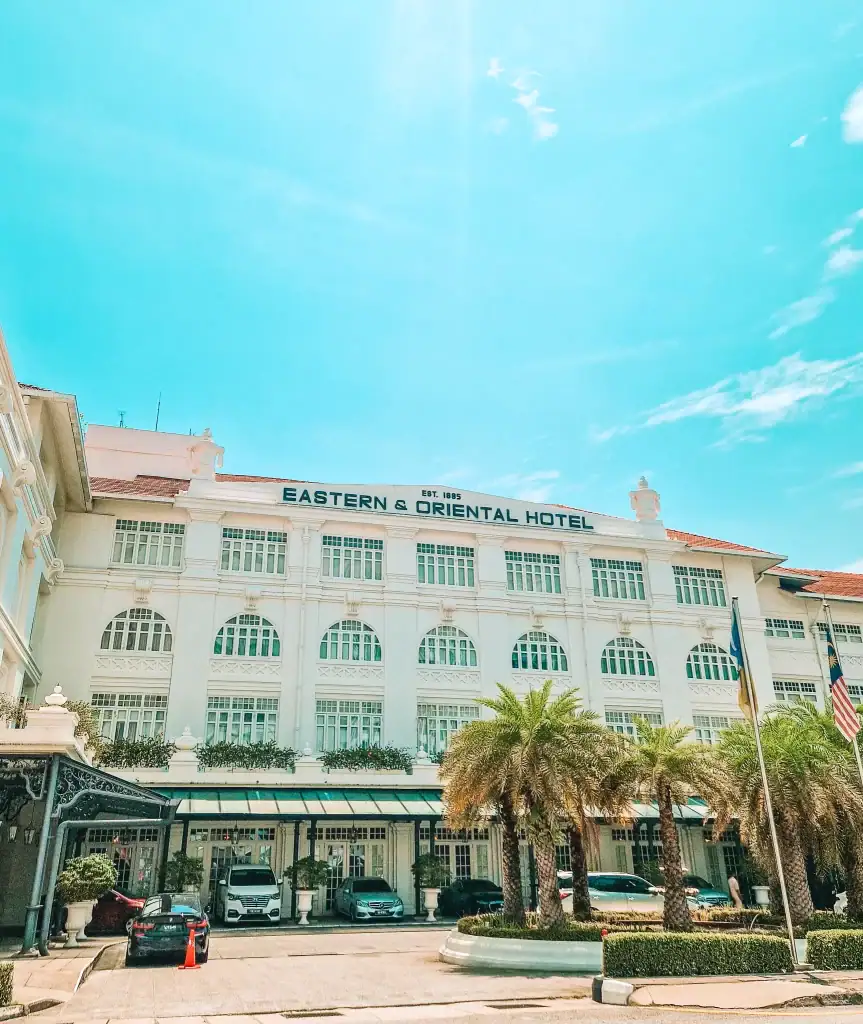 Grand colonial-style façade of the Eastern & Oriental Hotel in Penang, Malaysia, with arched windows, white exterior, and a red-tiled roof under a bright blue sky. Palm trees and parked cars line the hotel entrance, emphasizing its luxurious and historic ambiance.
