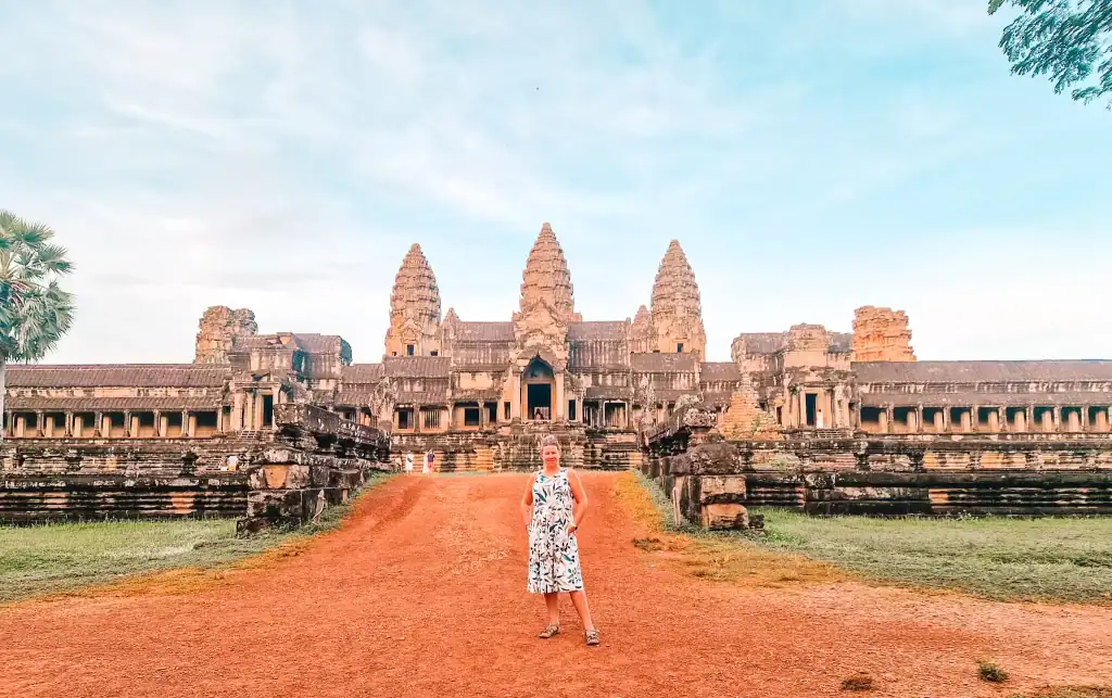 Em stands confidently on the red dirt path leading to Angkor Wat in Cambodia, with the ancient temple’s iconic stone towers rising behind her under a soft blue sky. The scene captures the grandeur of the historic site and the scale of its intricate architecture.