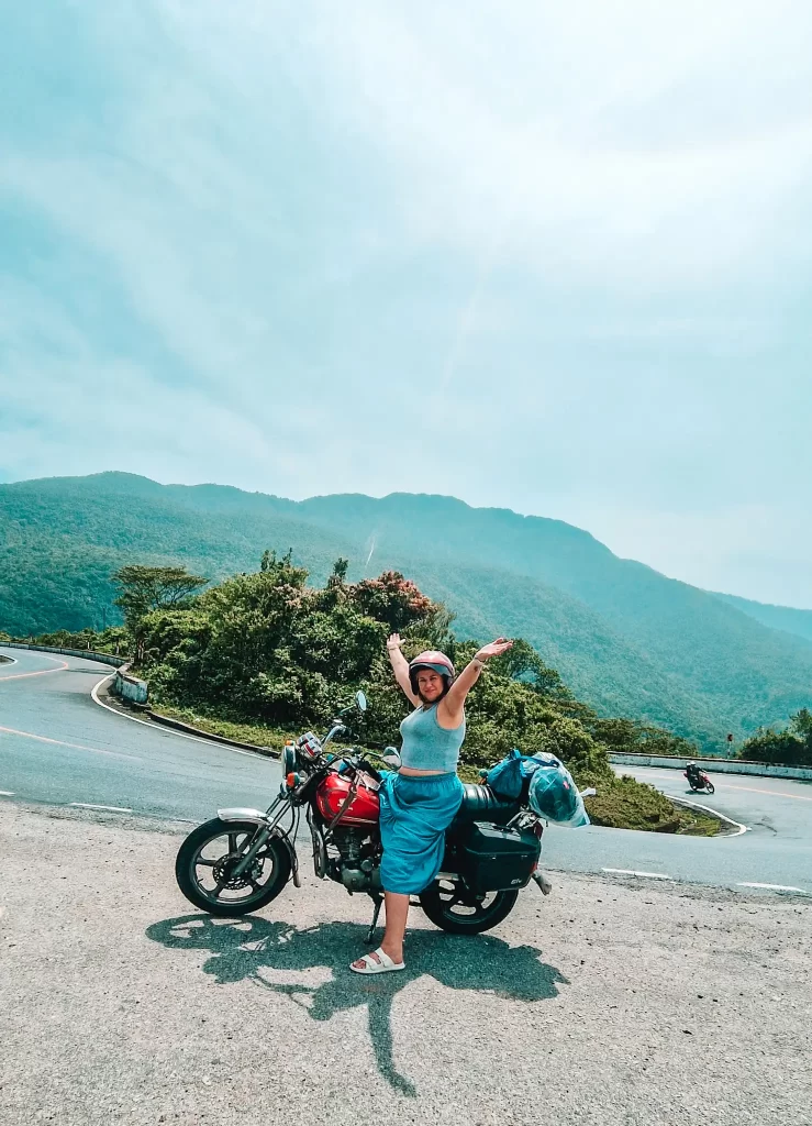 A smiling woman poses with arms raised beside a packed red motorbike on the Hai Van Pass, Vietnam. Behind her, the curving mountain road and lush green hills stretch under a bright blue sky.