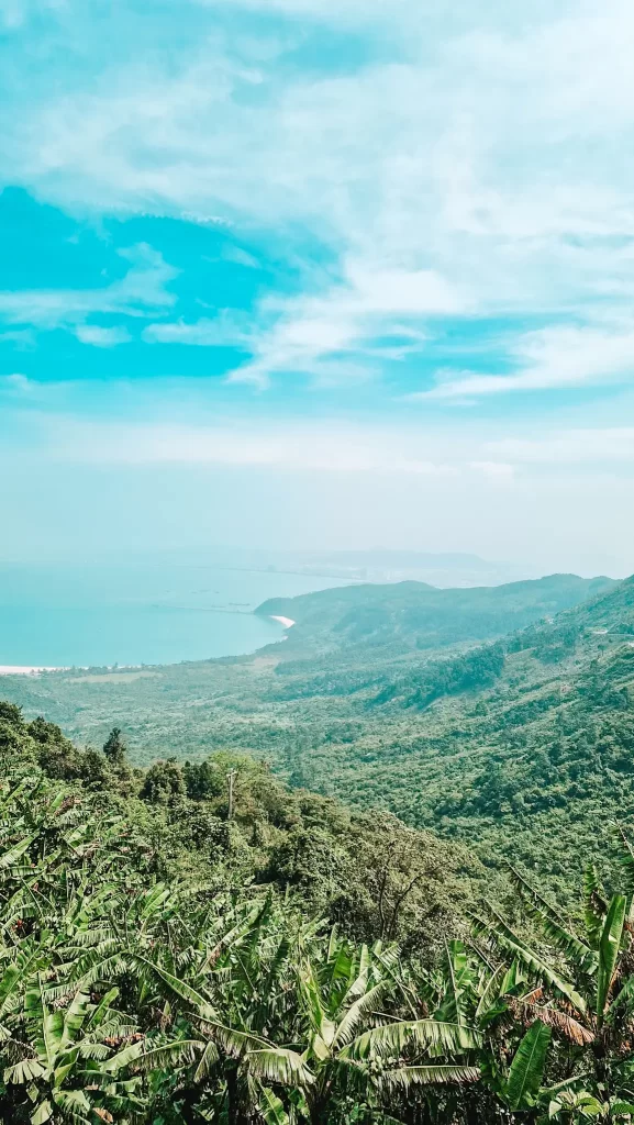 Panoramic view of a vibrant green mountainous landscape sloping down to a calm blue coastline, with a bright sky streaked with wispy clouds above.