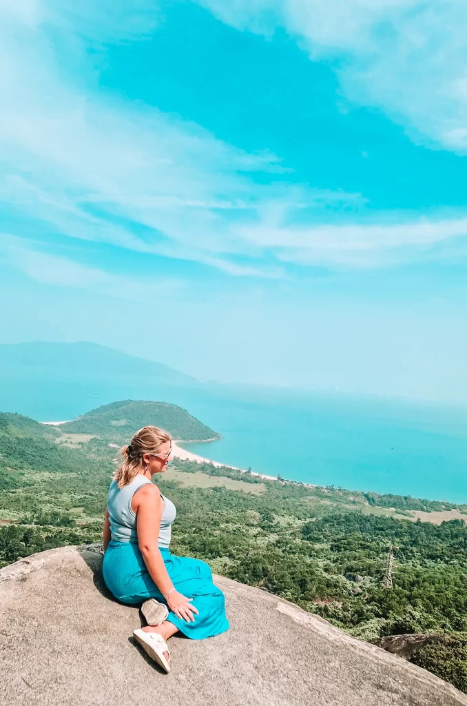 A woman in a blue skirt and tank top sits on a large rock overlooking sweeping views of forested hills and the ocean — a must-visit viewpoint in any Hai Van Pass guide.