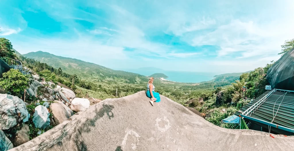 A woman ina blue skirt and a sleeveless top sits on a large rock, looking out over a sweeping green valley and distant ocean under a clear blue sky, with scattered boulders and a viewing deck nearby.
