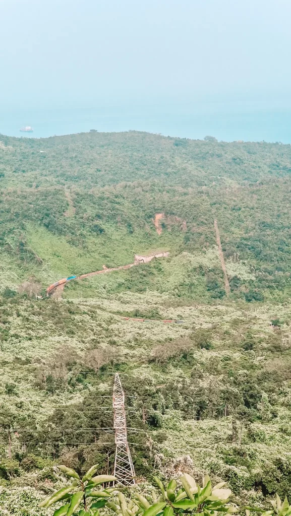 A panoramic view of a green, forested mountain with a train winding through a tunnel and a tall power line tower in the foreground. In the distance, the hazy blue sea meets the sky.