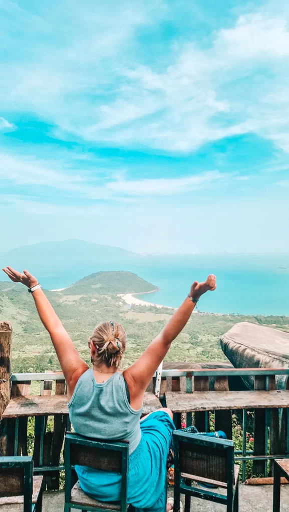 A woman with blonde hair in a bun, wearing a gray tank top and blue skirt, sits with arms raised in joy on a wooden deck overlooking a lush green island and turquoise sea, under a bright blue sky with wispy clouds.
