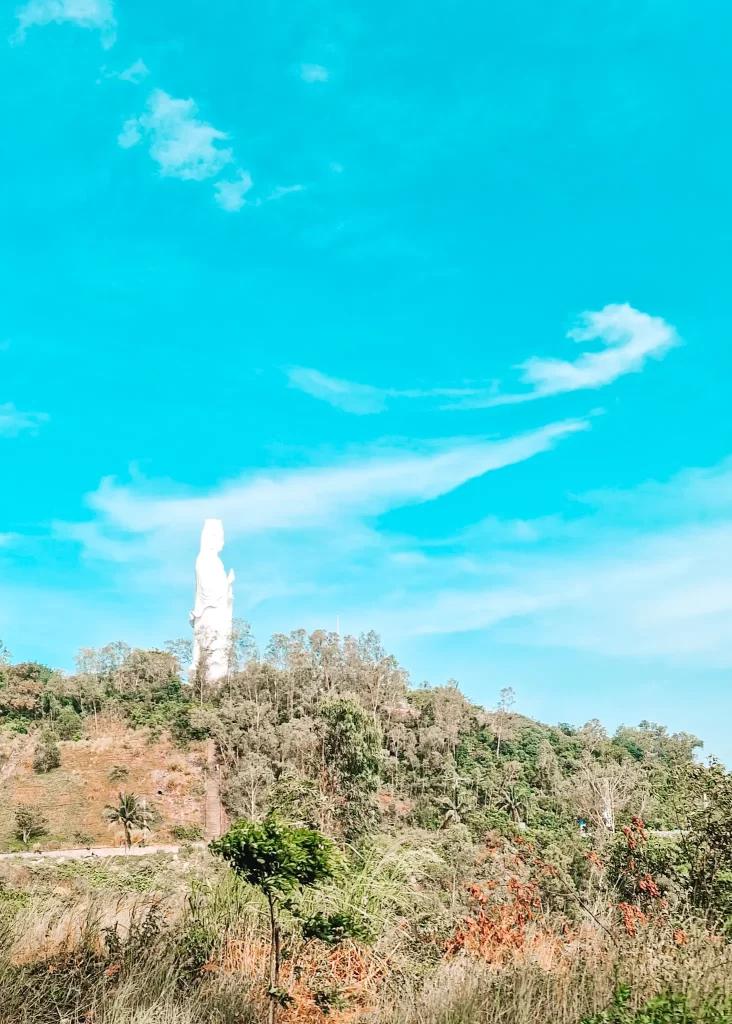  The tall white statue of Lady Buddha rises above the trees on a hill under a bright blue sky — a spiritual and photogenic stop featured in a detailed Hai Van Pass guide.
