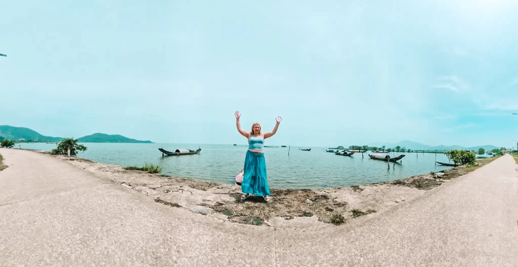 A woman in a blue outfit stands cheerfully with raised arms on a narrow concrete path flanked by water, with traditional Vietnamese fishing boats and distant green mountains in the background — an off-the-beaten-path stop on a Hai Van Pass guide.