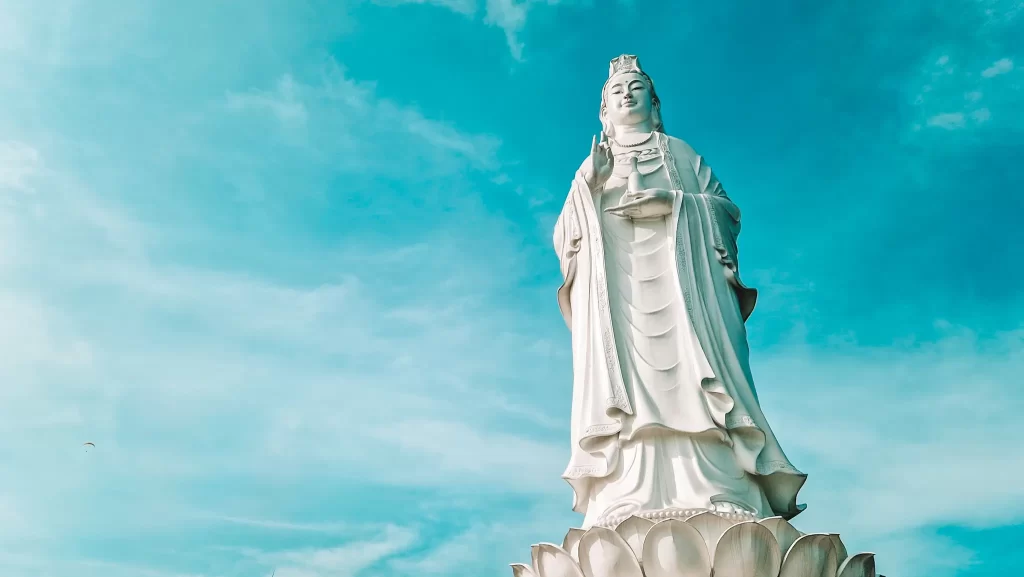  A towering white statue of the Bodhisattva Guanyin (Goddess of Mercy) stands against a bright blue sky with light clouds. The statue rests on a lotus pedestal and holds a small vase in one hand.