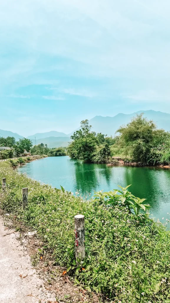 peaceful, emerald-green river winds through lush vegetation and wildflowers, with misty mountains rising in the distance — a scenic detour along the Hai Van Pass guide route.