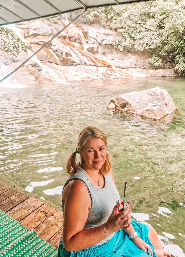 A woman in a grey tank top and blue skirt sits on a wooden deck by a natural pool, smiling while holding a drink, with a waterfall and rock in the serene forested backdrop.