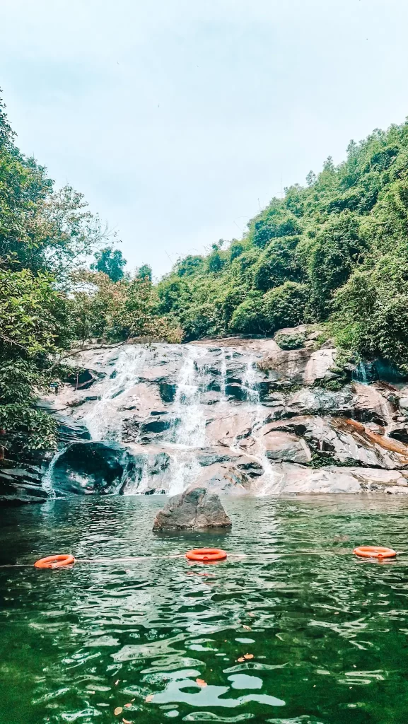Scenic waterfall cascading over smooth rocks into a green natural pool, surrounded by dense jungle; three orange life rings float in the foreground.
