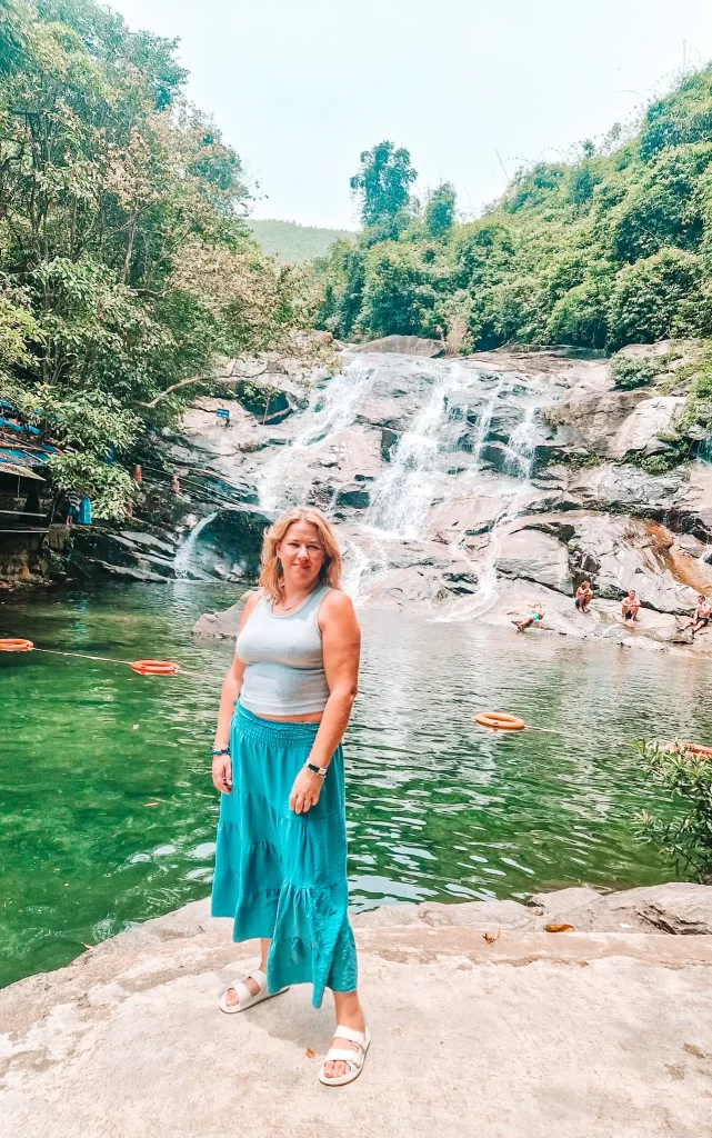 A woman in a light blue tank top and bright turquoise skirt stands on a rock ledge in front of a natural waterfall cascading into a clear green pool, surrounded by lush jungle. Other people are seen sitting on rocks and floating in the water.