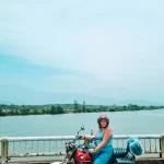 A smiling woman in a blue outfit and helmet straddles a red motorcycle parked on a bridge, with a scenic river, distant forest, and hazy mountains behind her under a clear sky.