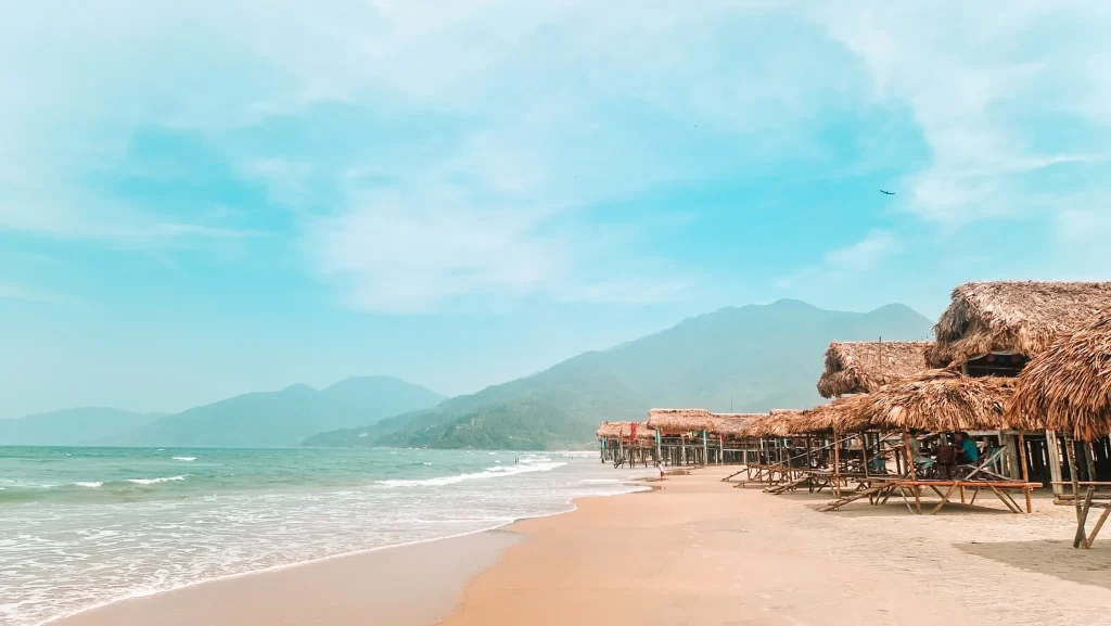 A quiet tropical beach lined with rustic bamboo and thatched huts, with gentle waves lapping at the shore and hazy mountains rising in the background.