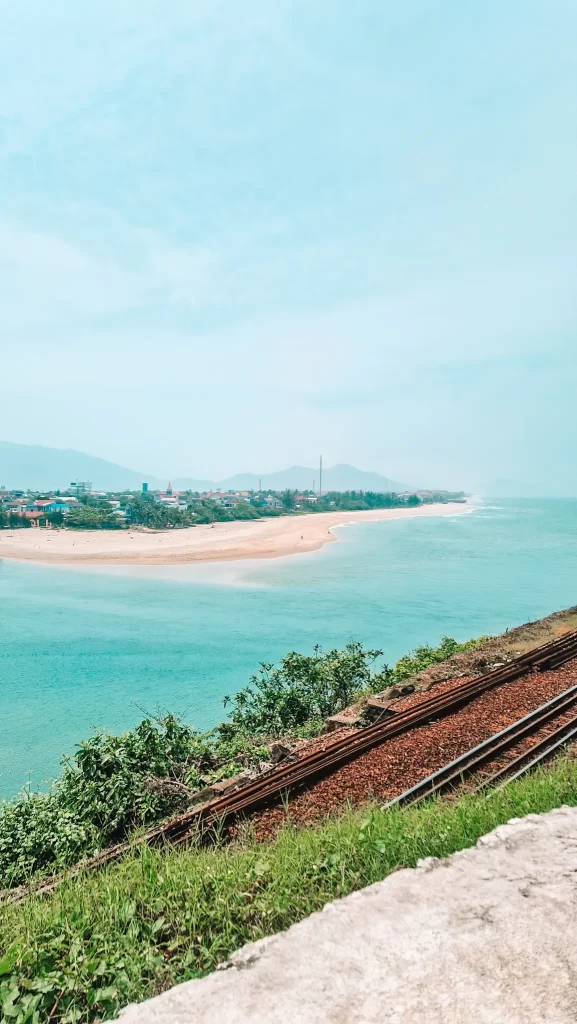 A scenic coastal view from the Hai Van Pass in Vietnam, showing turquoise waters meeting a sandy beach, with a small town and distant mountains in the background. In the foreground, railway tracks curve along the green hillside.