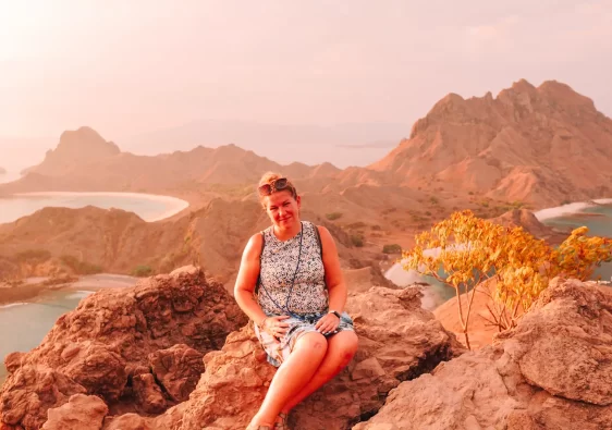 A solo female traveller sits on a rocky cliff at sunset, overlooking the rugged coastlines and curved bays of Komodo Island, Indonesia.