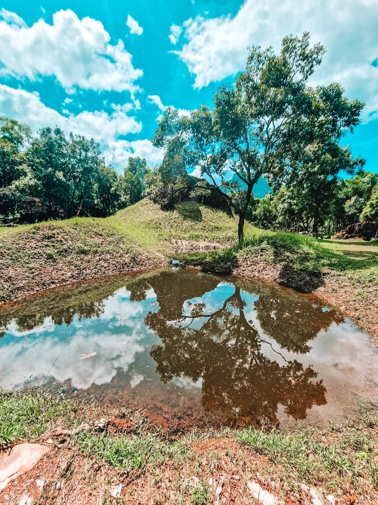 A bomb crater filled with still water reflecting the sky and trees, located at the historic Mỹ Sơn Sanctuary—an evocative reminder of war history often seen during day trips from Hoi An.