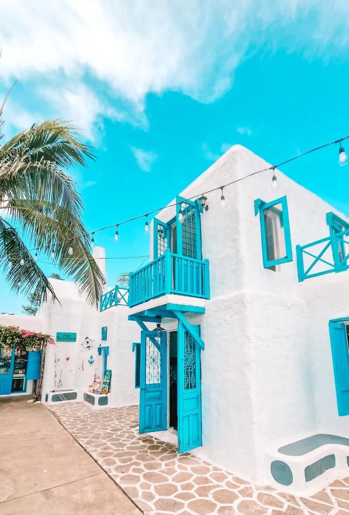  A charming white stucco building with vivid blue doors, windows, and a small balcony stands under a blue sky. Decorative lights hang overhead, and palm leaves and nautical-themed decor add to the Mediterranean-inspired vibe.