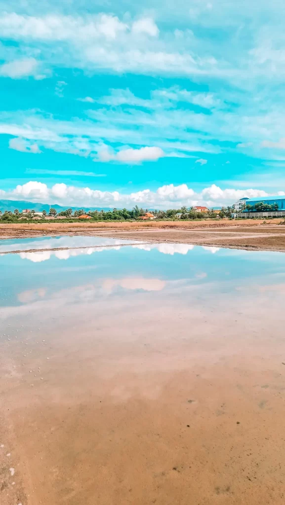 Still salt flats in Kampot reflect the vivid blue sky and scattered white clouds, with distant homes and palm trees lining the horizon. Exploring the salt flats is a scenic and culturally rich experience during 2 days in Kampot.