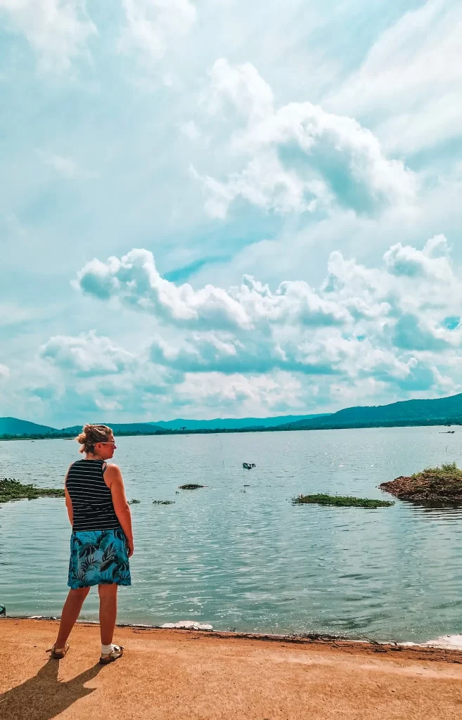 A woman in a striped tank top and tropical-print skirt gazes out over the calm waters of the Secret Lake in Kampot, with clouds drifting across the sky and mountains in the distance. A peaceful detour to include during 2 days in Kampot.
