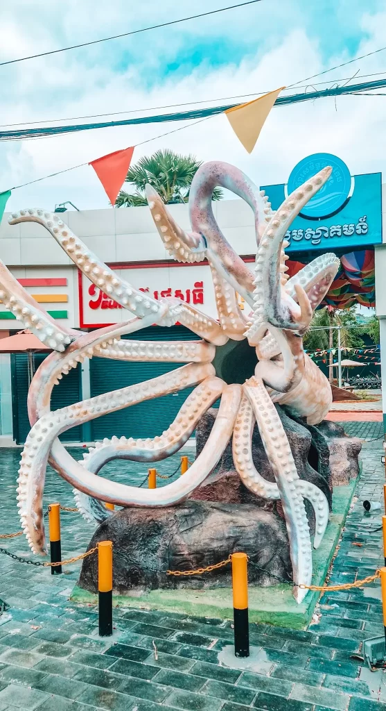 A massive white octopus sculpture with curling tentacles stands in a plaza near Kep’s waterfront, surrounded by colorful flags and shops. A quirky coastal stop worth seeing near Kampot during 2 days in Kampot.
