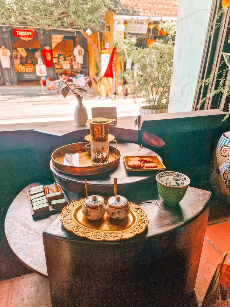 A cozy tea setup inside a café, featuring ornate gold-trimmed teacups and a traditional drip coffee filter, with a view of Hoi An street shops outside.