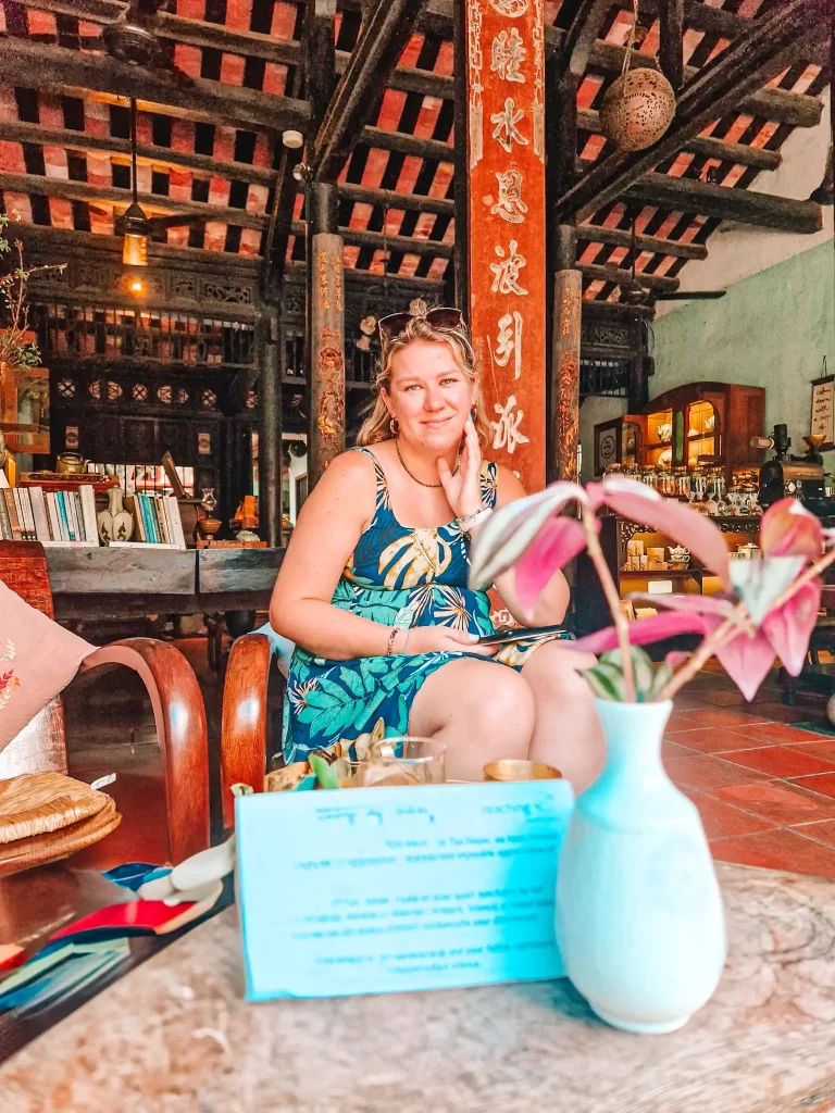 A woman in a tropical-print dress sits smiling inside a traditional Vietnamese café, with wooden beams and red columns inscribed with golden Vietnamese text behind her.