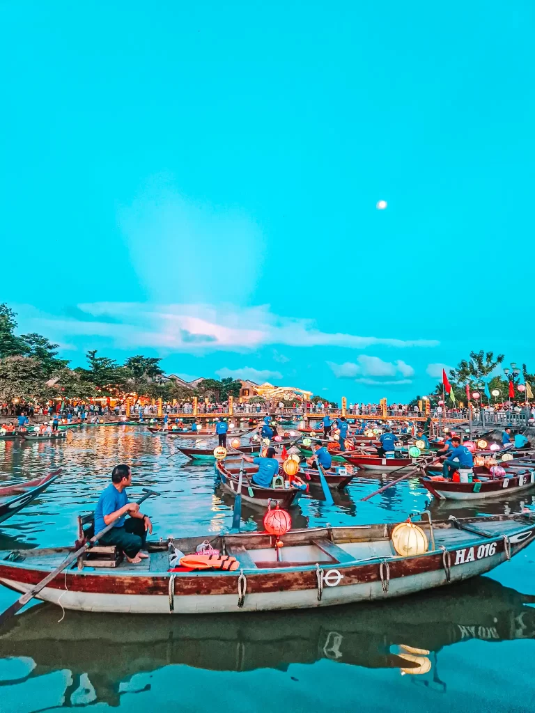 A vibrant river scene at sunset with wooden boats filled with people and colorful lanterns, reflecting on the water as crowds gather along the riverbank under a glowing blue sky.
