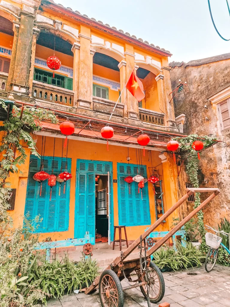 A rustic two-story yellow house with turquoise shutters, decorated with red lanterns and a Vietnamese flag, and an old wooden handcart parked in front amid lush plants.