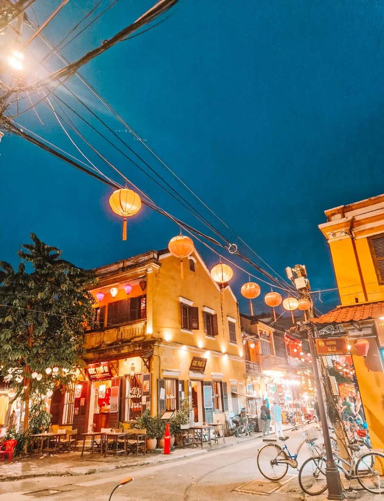 A bustling night street in Hoi An, Vietnam, glowing with hanging orange lanterns and string lights, with cafes and shops open, bicycles parked, and people strolling.