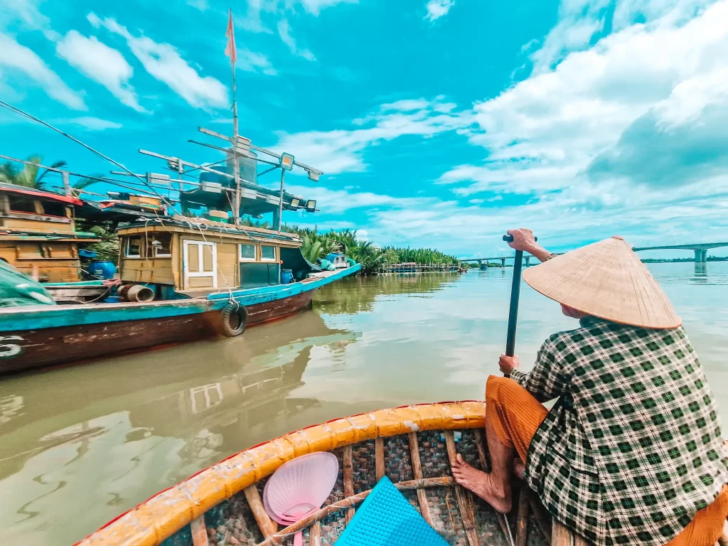A person wearing an iconic conical hat rows a circular basket boat past rustic fishing boats on a calm river, with a bridge and dense greenery in the distance beneath a partly cloudy sky.