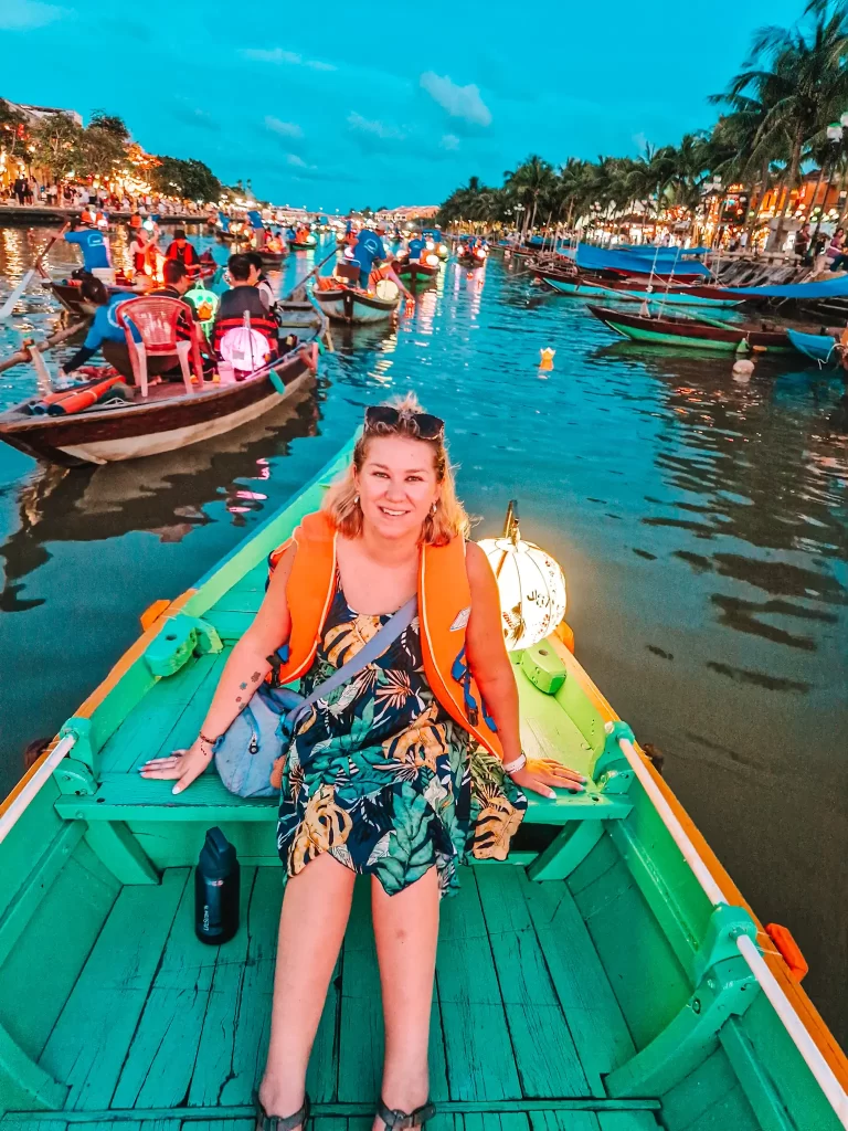 smiling woman in a colorful dress and orange life jacket sits on a green wooden boat surrounded by lantern-lit rowboats during sunset in Hoi An. This magical boat ride experience is one of the best things to do in Hoi An.