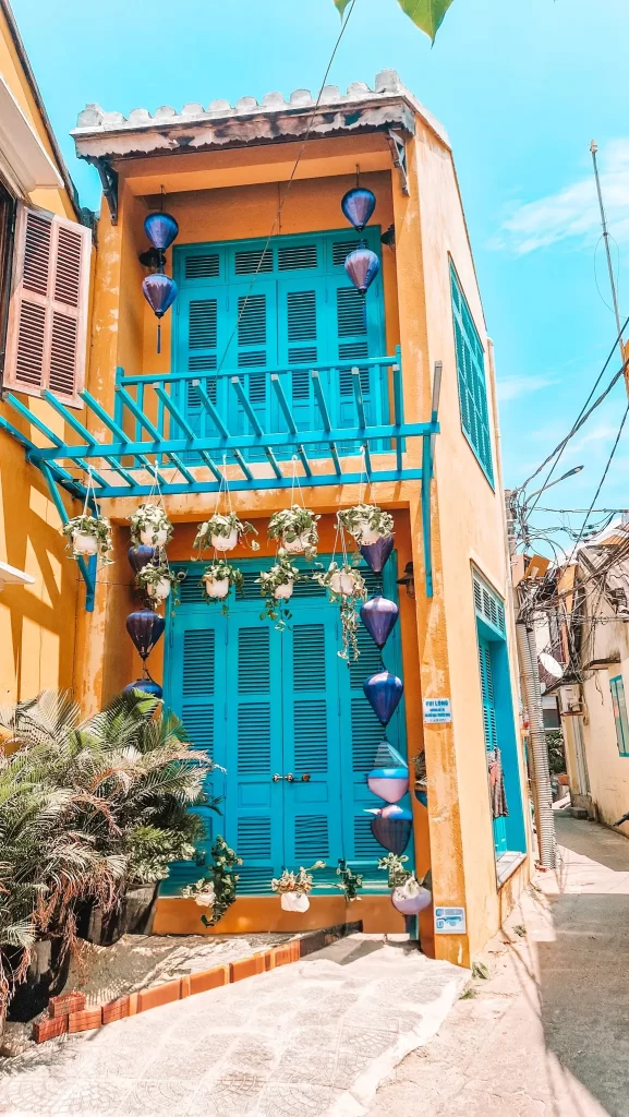 A charming narrow house painted in golden yellow with vivid turquoise shutters and doors, decorated with hanging potted plants and blue lanterns under a sunny sky.
