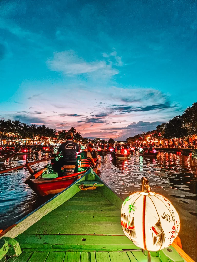A view from a green wooden boat cruising through Hoi An’s lantern-lit river at dusk, with dozens of boats and glowing lanterns filling the waterway. Enjoying a traditional boat ride at sunset is among the best things to do in Hoi An.
