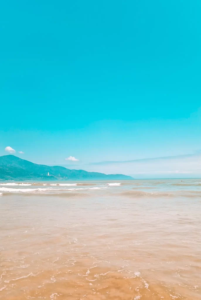 A serene beach scene with gentle waves and distant green mountains under a bright blue sky in Hoi An, Vietnam. Relaxing at the beach is definitely one of the best things to do in Hoi An for nature lovers.