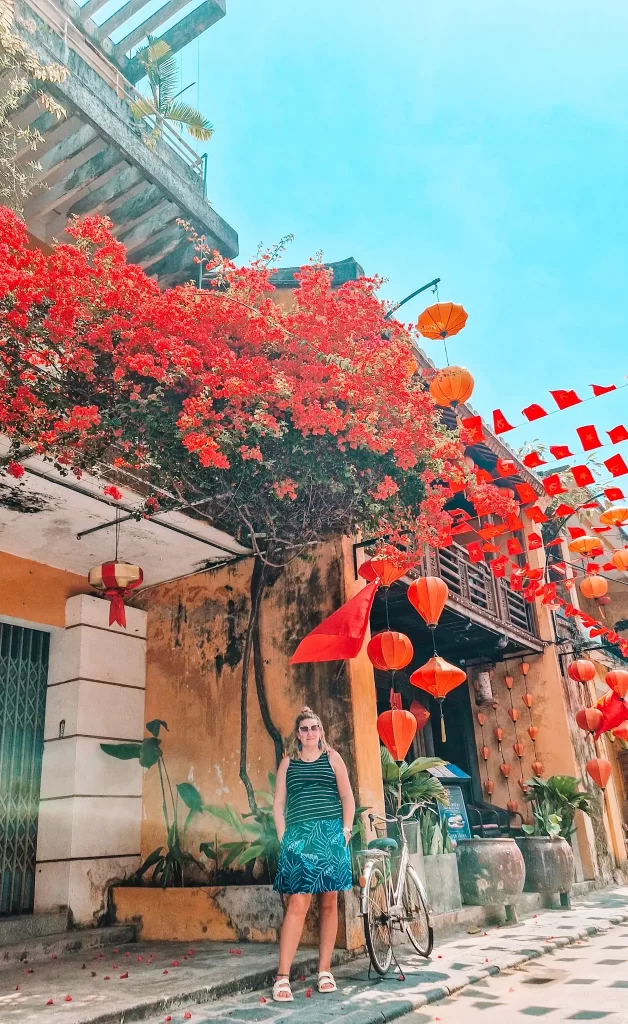 A woman in a striped dress stands next to a white bicycle on a charming Hoi An street, under vibrant red bougainvillea flowers and a canopy of hanging red lanterns and flags.