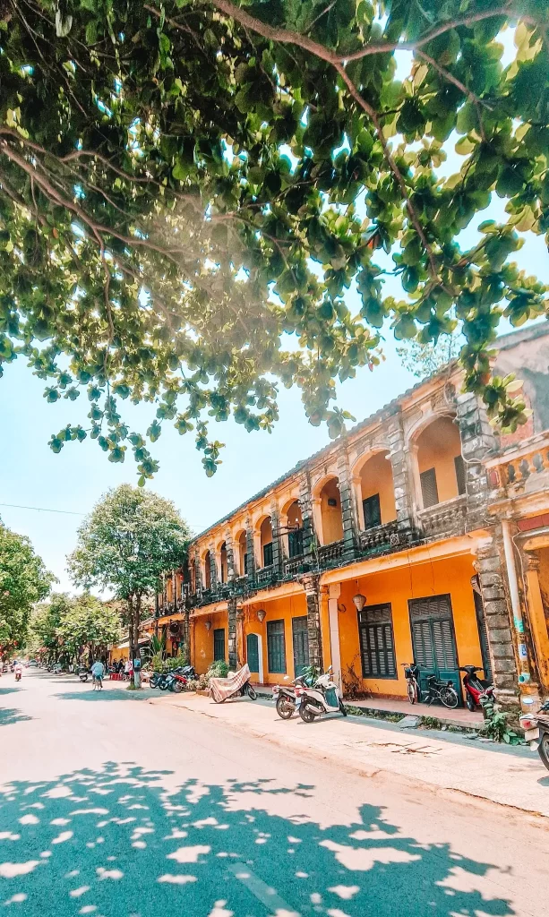 A quiet street shaded by leafy trees, with a row of two-story colonial-style yellow buildings featuring arched balconies and parked motorbikes in front.