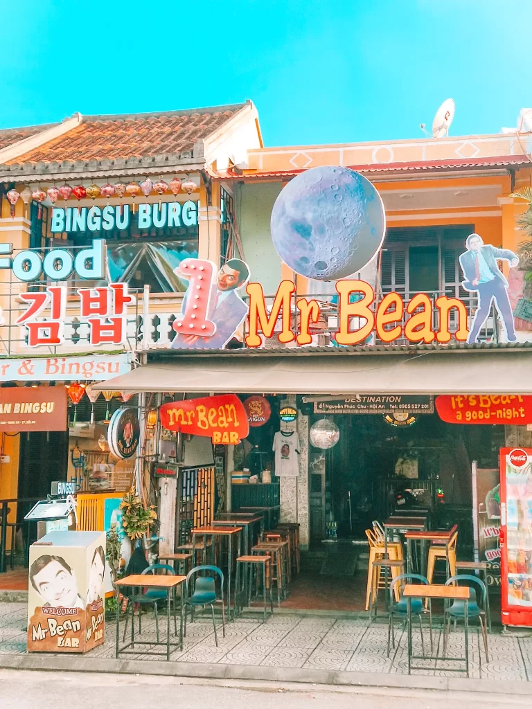 Front view of a quirky bar in Hoi An decorated with Mr. Bean images, Korean signage, a giant moon cutout, and rows of tall stools under a bold sign that reads “Mr Bean Bar.”