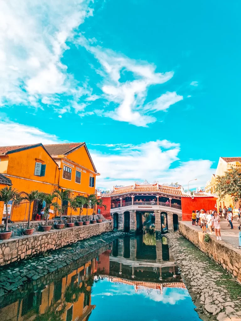 A scenic view of the iconic Japanese Covered Bridge in Hoi An, Vietnam, reflected in a narrow canal flanked by mustard-yellow buildings and groups of tourists.