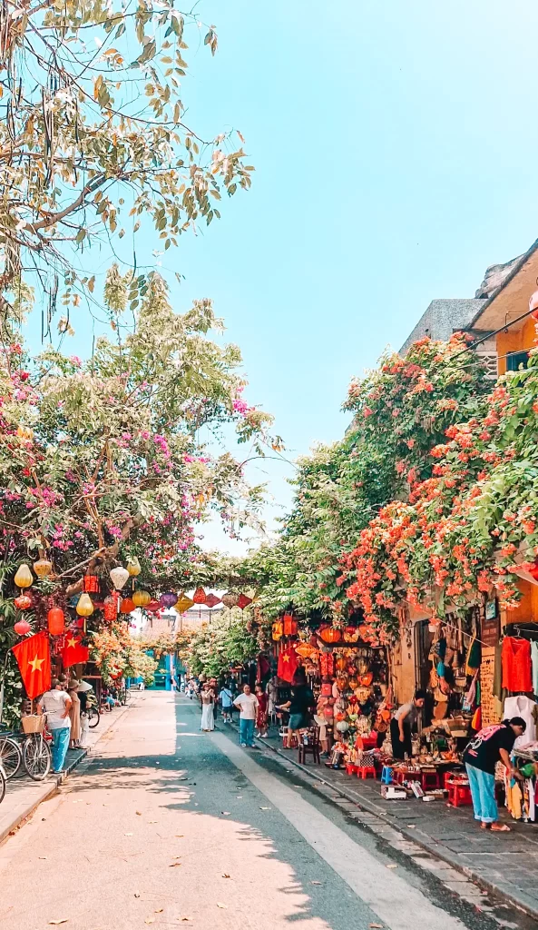 A lively street scene in a Vietnamese market, with colorful lanterns hanging overhead, lush flowering vines spilling over buildings, and vendors selling crafts and souvenirs along the sidewalk as people stroll through under a clear blue sky.