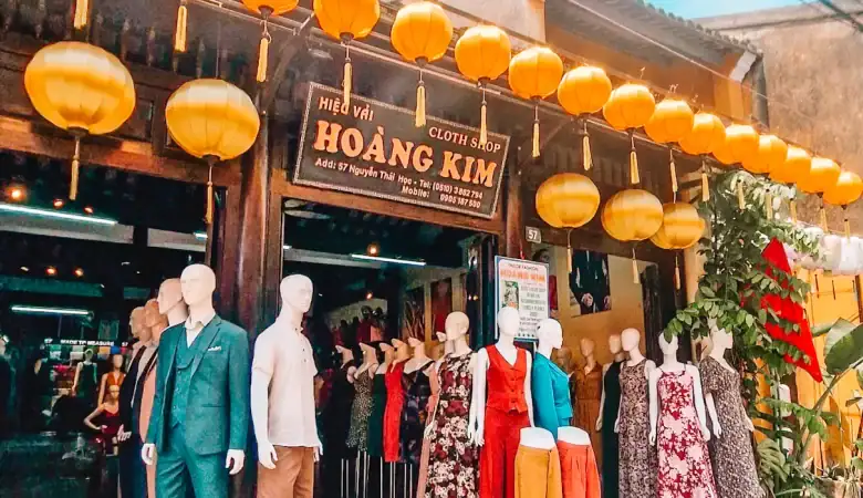 The Hoang Kim tailor shop in Hoi An, showcasing brightly colored clothing on mannequins and rows of yellow lanterns strung above the entrance.