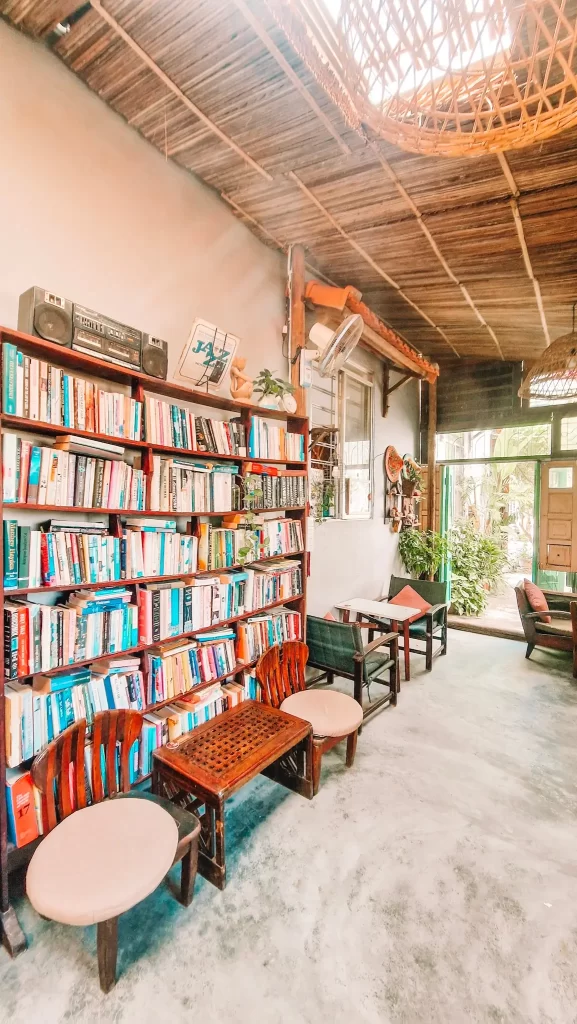 A cozy indoor corner featuring a tall wooden bookshelf filled with colorful books, vintage decor like a boombox and vinyl records, low wooden chairs with round cushions, and a rustic ceiling made of bamboo slats letting in sunlight.