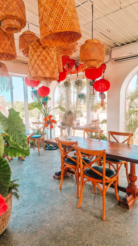  A cozy café interior with wooden chairs and tables, lush potted plants, and hanging rattan and red lantern-style lights, bathed in natural light from large arched windows.
