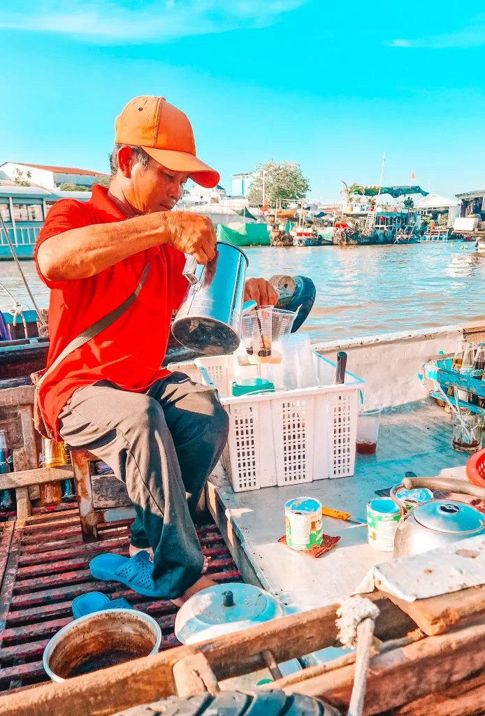 A vendor in a red shirt and cap pours coffee from a metal pot into plastic cups on a boat at Cai Rang floating market, with bustling boats and riverside stalls in the background. A must-see local experience during 4 days in the Mekong Delta.