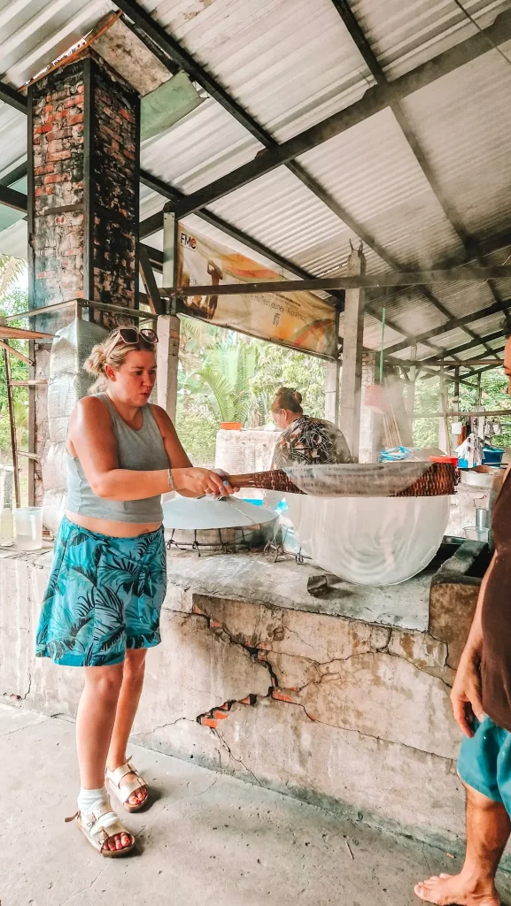 Emr uses a bamboo frame to lift freshly steamed rice noodles from a hot surface inside a traditional noodle factory in the Mekong Delta. Learning how local rice noodles are made is a hands-on highlight of spending 4 days in the Mekong Delta.