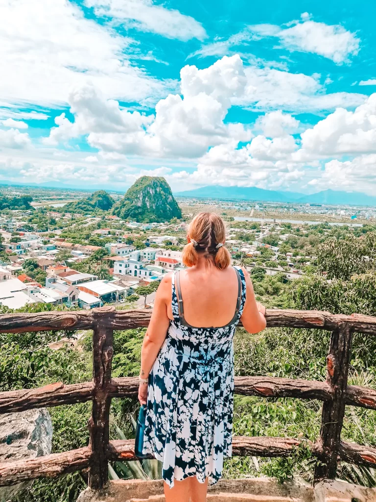 A woman in a black-and-white floral dress stands at a rustic wooden railing, looking out over a sprawling town with a lush, green mountain in the background under a vibrant blue sky dotted with fluffy clouds.