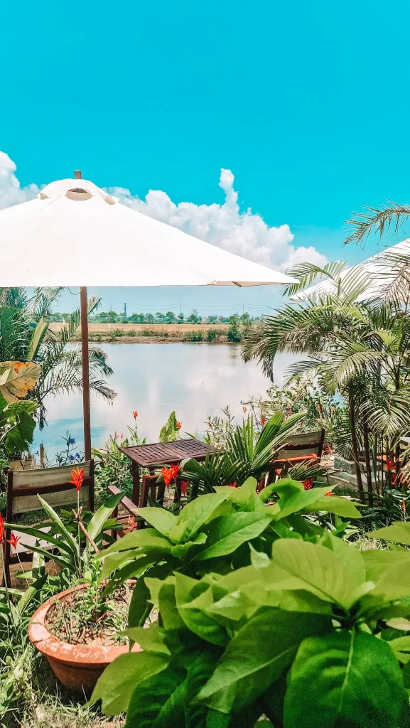 A cozy outdoor café by a calm river features wooden tables shaded by large white umbrellas, surrounded by tropical plants and bright red flowers, all under a clear blue sky.